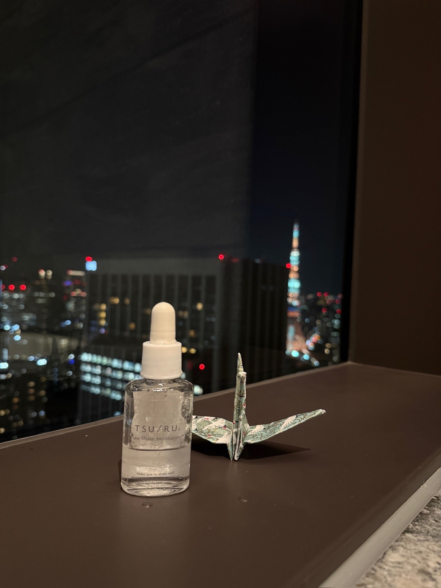 夜景と折り鶴とともに置かれたTSURU Pure Shake Moisturizer